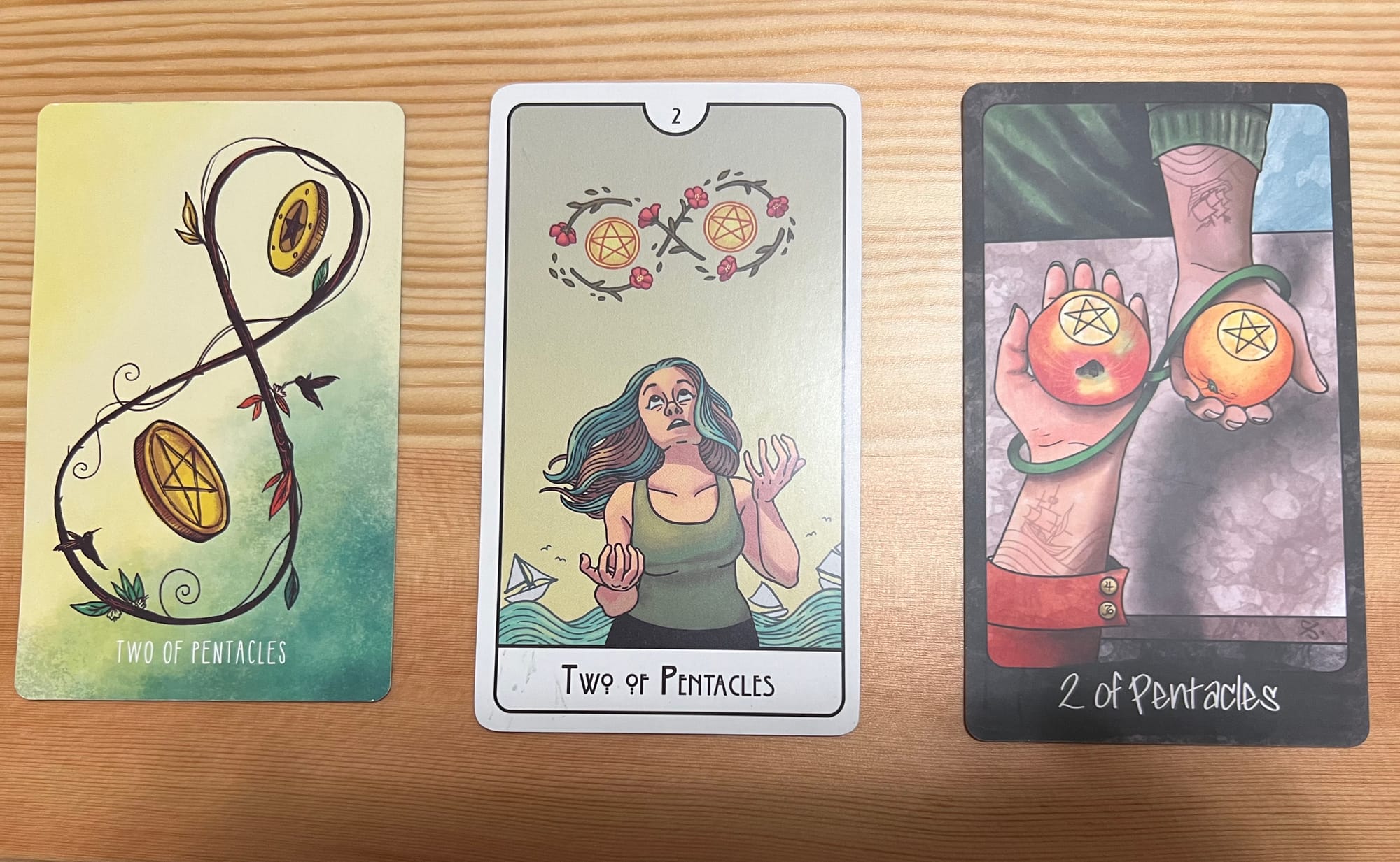 Three examples of the Two of Pentacles card from different tarot decks. The first, from the Spacious Tarot, shows two coins floating within the figure-eight of a vine. The second, from the This Might Hurt Tarot, shows a light-skinned woman with brown and blue hair in a green tank top and black pants juggling two coins within a figure-eight of flowers, while sailboats traverse waves in the background. The third, from the Out of Hand Tarot, shows the left hands of two figures extended across a table, one with a red sleeve and one with a green sleeve; each hand holds an orange with a pentacle carved in it, and their wrists are wrapped in a green band twisted in a figure-eight.